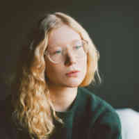 Image of woman wearing black sweatshirt and glasses looking forward-right, sitting in chair with white curtain and black wall in background