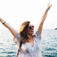 Woman with arms raised over head, sunglasses on, smiling, with ocean in the backgronud