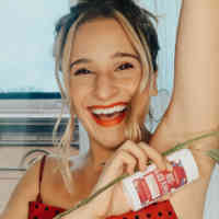 Woman smiling with blonde hair in a bun wearing a red and black polka dot tank top lifting her arm up and holding a rose and a Rose+Black Pepper Hemp Seed Oil deodorant from Schmidt's