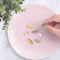 Pink plate with assortment of pill supplements on top with manicured hand picking up one of the pills with eucalyptus plant nearby on table with plate