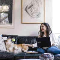 A woman with a laptop petting her dog on a sofa